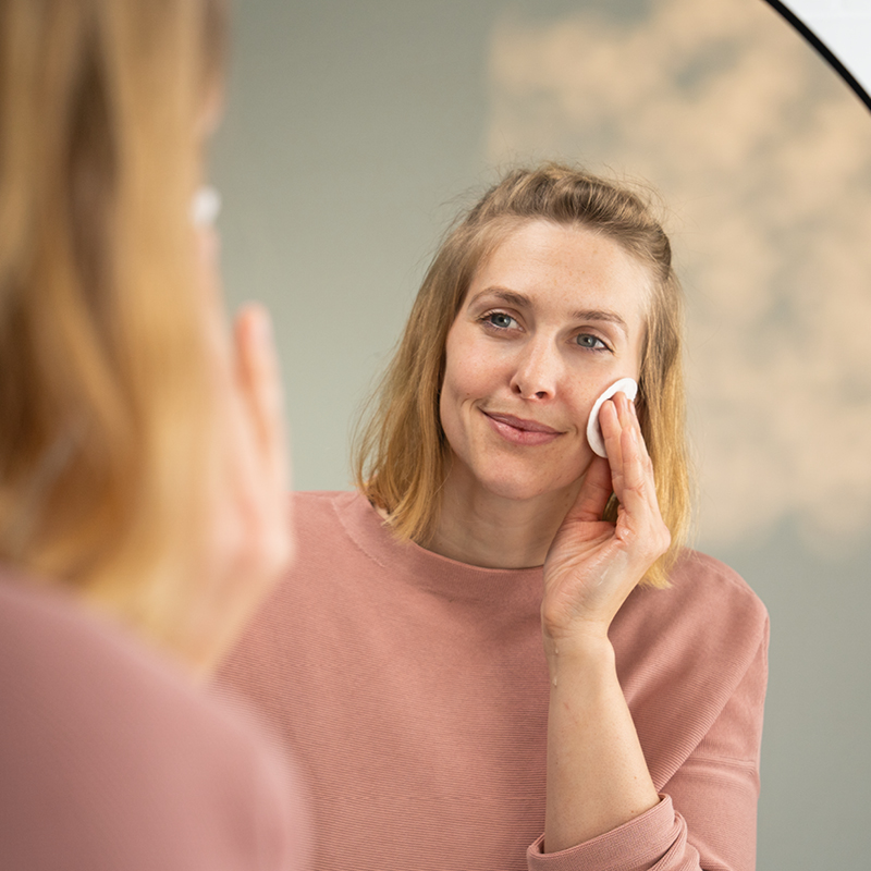 Vrouw brengt glimlachend voor een spiegel een product met een wattenschijfje aan op haar gezicht