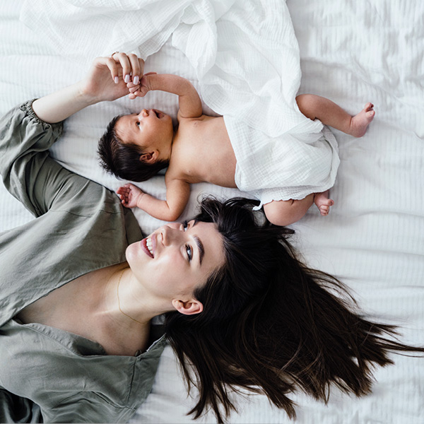 [Translate to Englisch:] Dunkelhaarige Mutter liegt neben ihrem neugeborenem Baby auf dem Bett